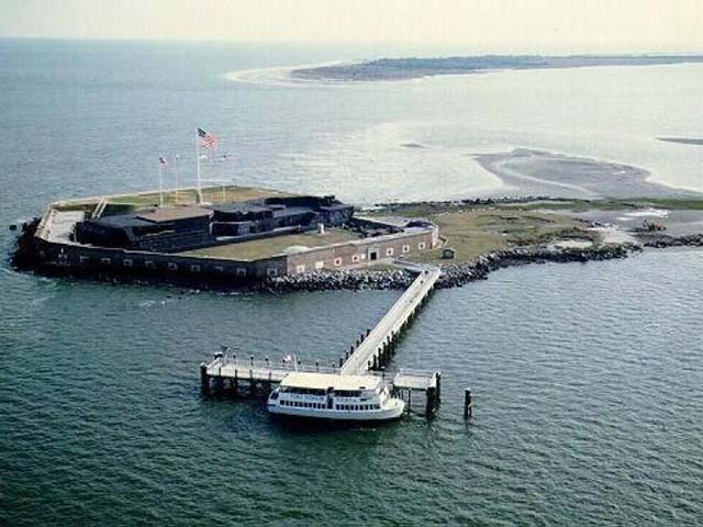 A five-sided brick fort with black buildings and flags inside the fort. The fort occupies about half of the island. A pier leads to a docked boat.